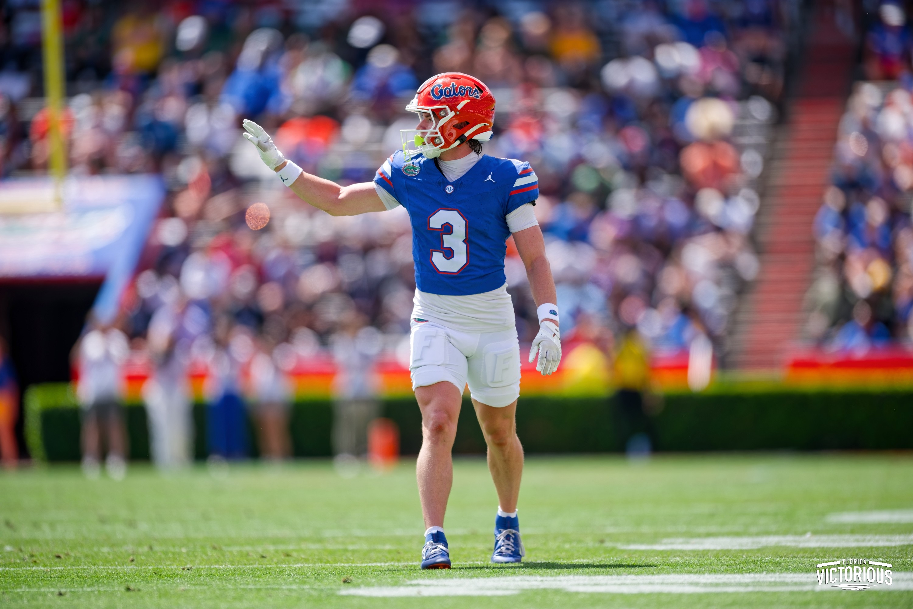 Bailey Stockton in a blue jersey and orange helmet, numbered 3, stands on a sunny football field, raising his left hand as he looks to the side. The crowd in the stadium is blurred in the background.