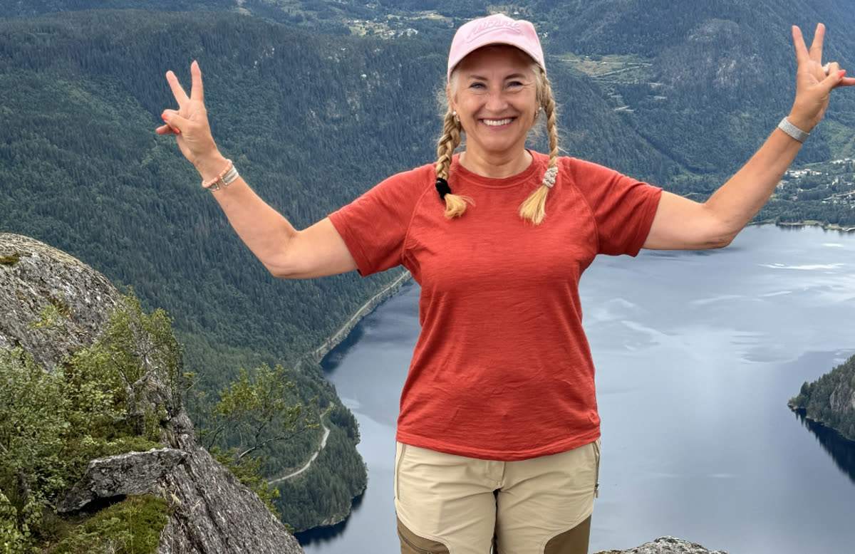 May Ruth på en fjelltopp med armene i været, med fjord og skog bak seg