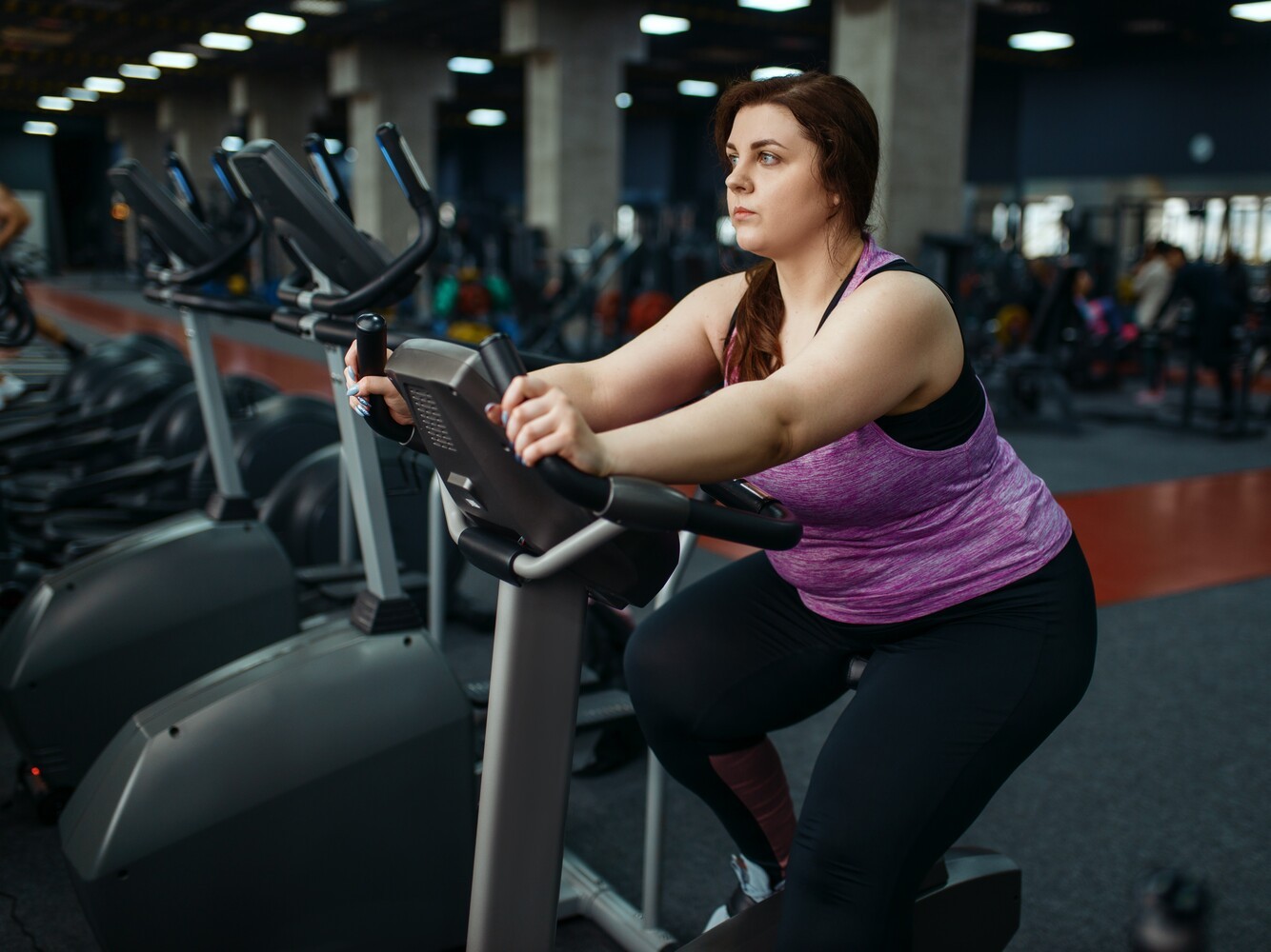 overweight woman using a stationary bike as part of her exercise routine at the gym before using a steam room for body composition