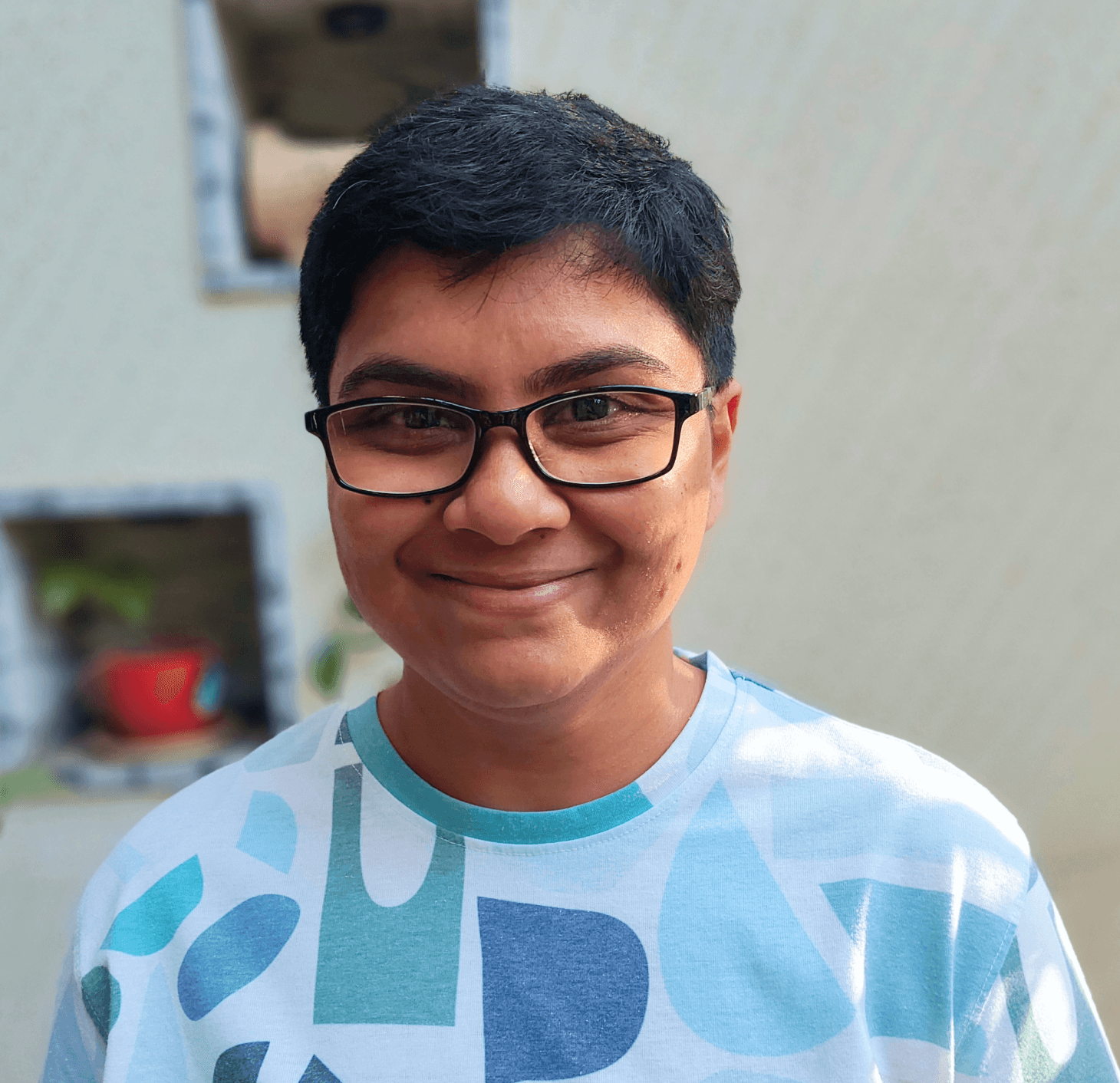 A picture of Jade — short black hair, rectangular black glasses, smiling at the camera. Wearing a t-shirt with white, blue and green organic shapes. Daytime light falling softly on the left side.