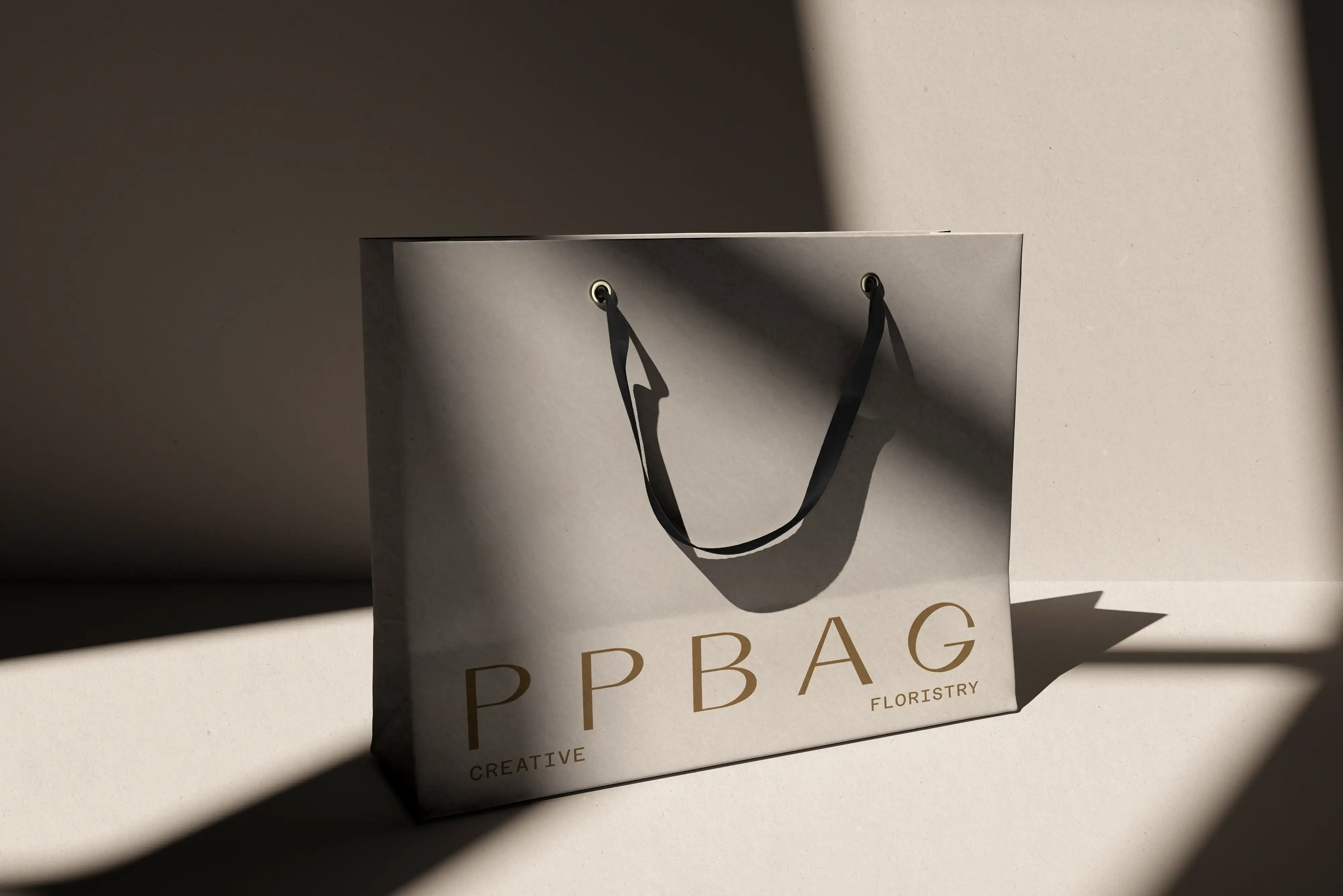 White paper shopping bag with black rope handles and a spaced-out wordmark in hard shadow