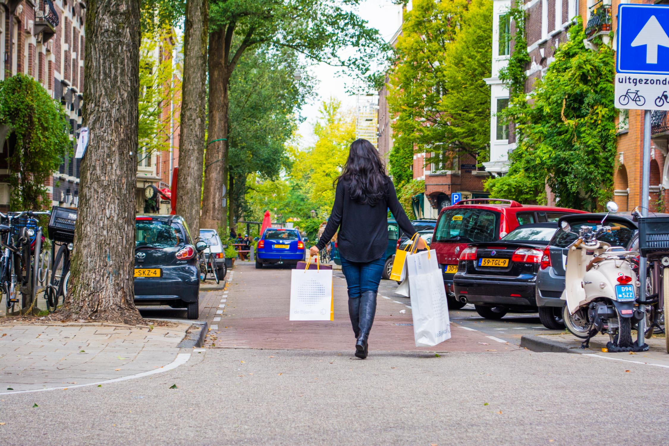 People walk down a tree-lined, paved thoroughfare under bright daylight, passing colorful buildings and parked vehicles.