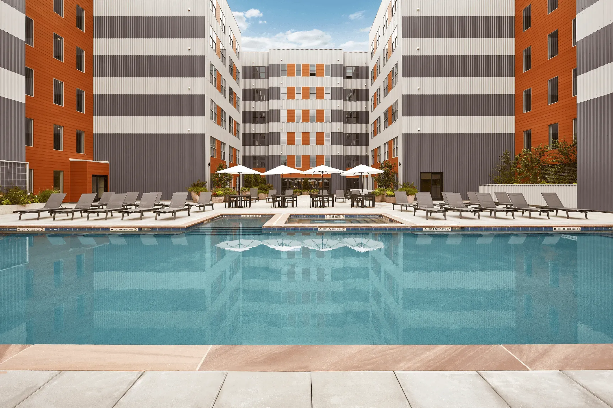 Modern apartment complex with a pool, lounge chairs, and landscaped areas, set against a clear blue sky.