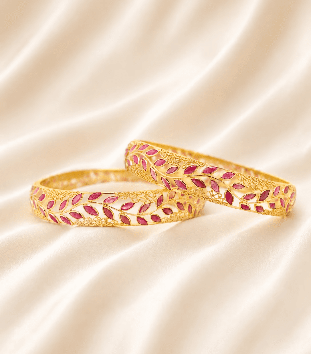 A pair of gold bangles with ruby leaf designs are displayed on a cream silk fabric.