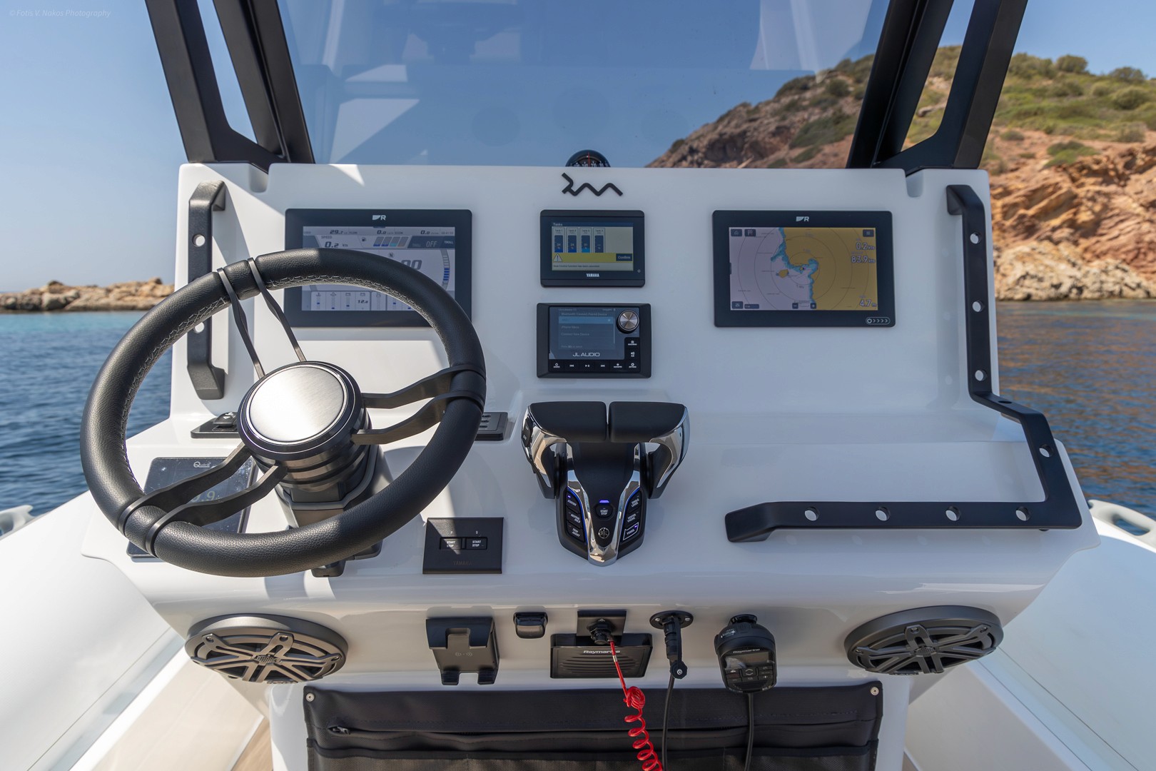 Modern yacht helm station with multiple navigation screens and controls, overlooking calm blue waters and rocky coastline in the Greek islands.