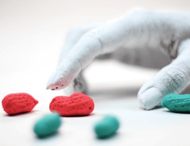 A hand with white paint is reaching toward three small, crocheted heart-shaped objects on a white surface. The hearts are red and teal/turquoise. The hand is positioned near the hearts, suggesting an action or interaction. The background is a plain white surface.