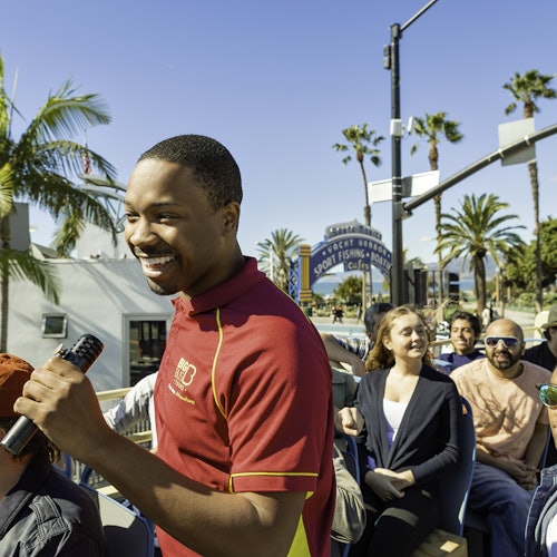 A smiling tour guide with a microphone leads a group of people on an open-top bus tour near a waterfront in sunny weather.