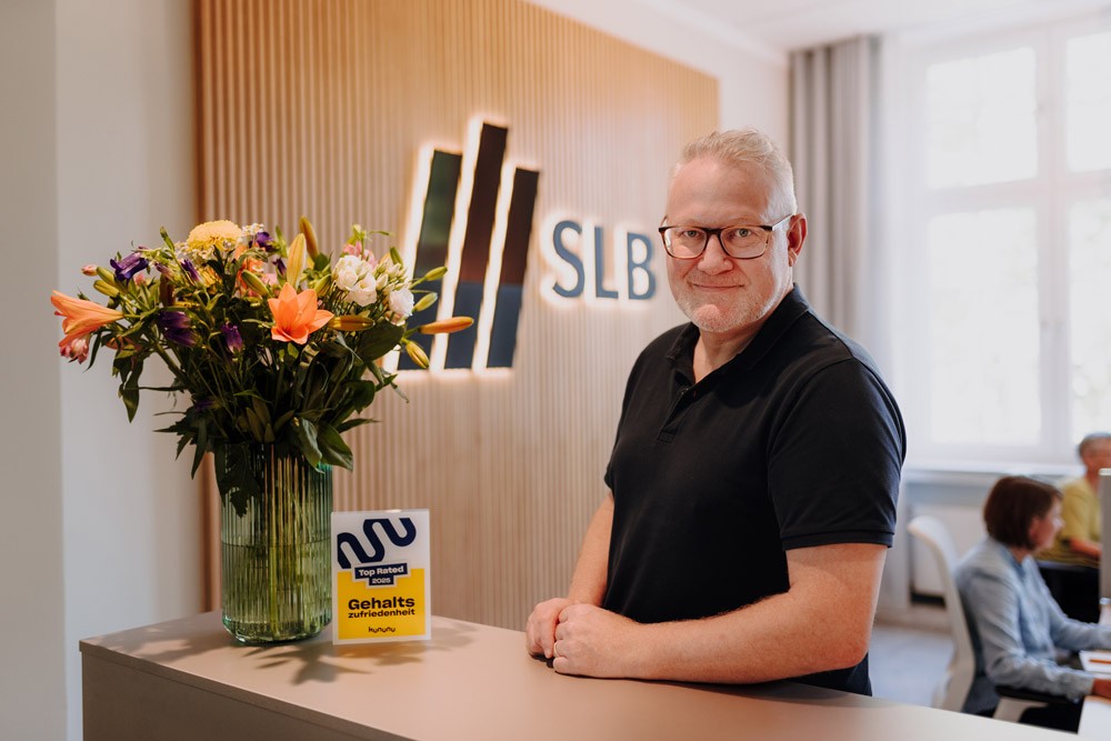 Mitarbeiter steht hinter dem Empfangstresen bei SLB, neben ihm ein großer Blumenstrauß und das SLB-Logo an der Wand.