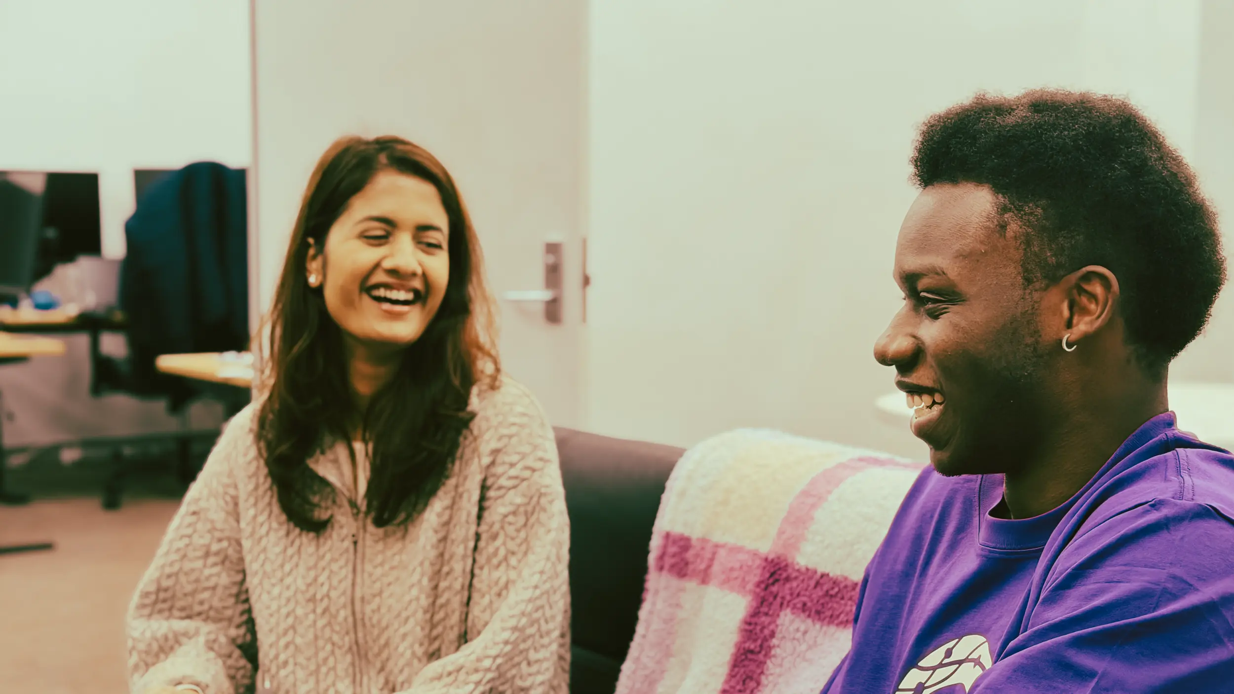 Two people are sitting on a couch in a casual office setting, engaging in a lively conversation and sharing a laugh, with computers seen in the background.