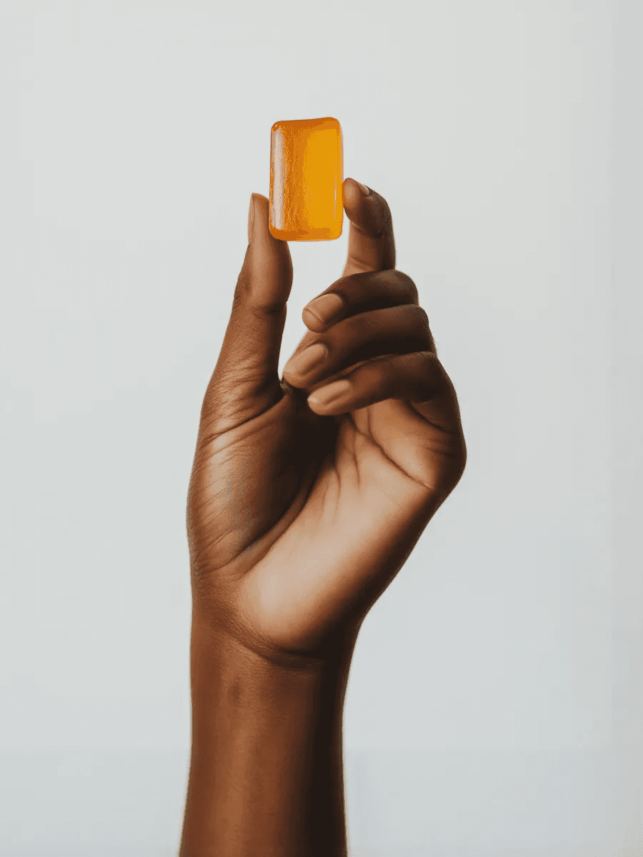 Hand holding up a translucent amber-colored gummy or supplement against a plain light background.
