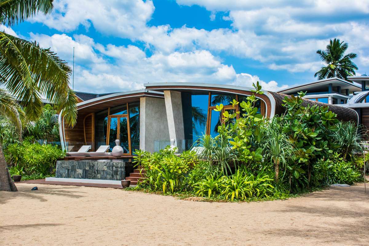 A modern, curved concrete villa with large glass windows sits nestled among tropical greenery and palm trees under a blue