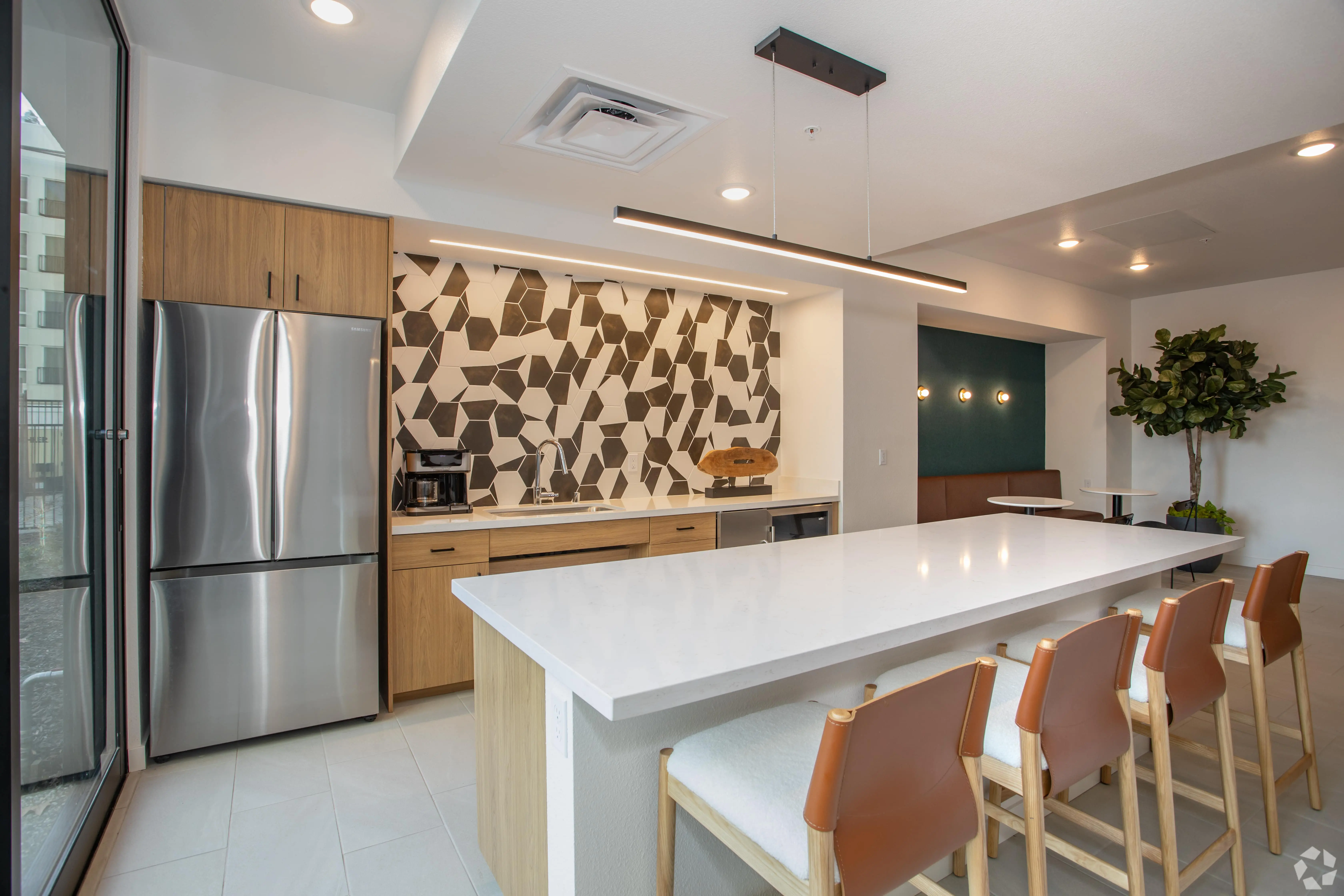 A modern kitchen in Midtown Sacramento with a white island countertop and four barstools featuring light-colored wood legs and leather seats. This sun-drenched living space boasts stainless steel appliances, light wood cabinetry, a geometric-patterned backsplash, a small dining area, and a potted plant.