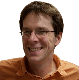 A man with short brown hair, glasses, and a warm smile is pictured indoors, wearing an orange shirt. The relaxed setting and natural smile give the image a friendly, approachable feel, ideal for portraying a professional in a casual environment.