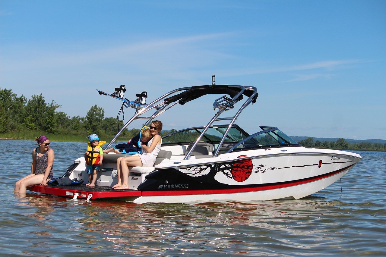 A family enjoys a sunny day on a sleek, white and red Four Winns motorboat floating on a calm lake, surrounded by lush greenery and clear blue skies.