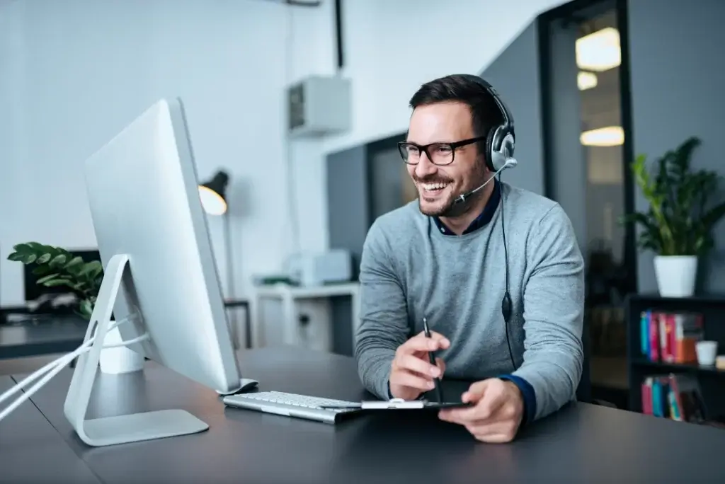 Image of a man sat at a desk, looking at a desktop screen, wearing a headset and smiling.