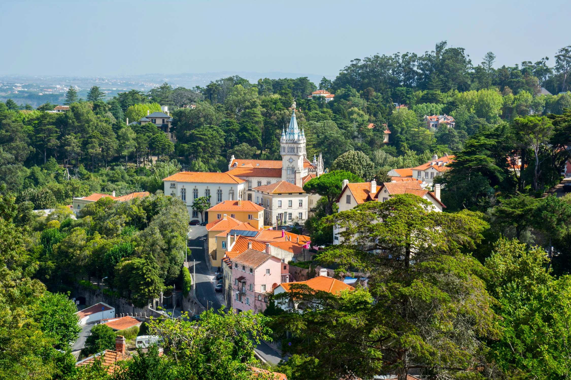 A high-angle view of a coastal village with terracotta-roofed houses and a white church tower nestled among green trees.