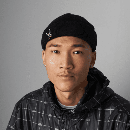 A young person wearing a black beanie adorned with a small silver emblem and a checkered shirt, looking directly at the camera with a calm expression.