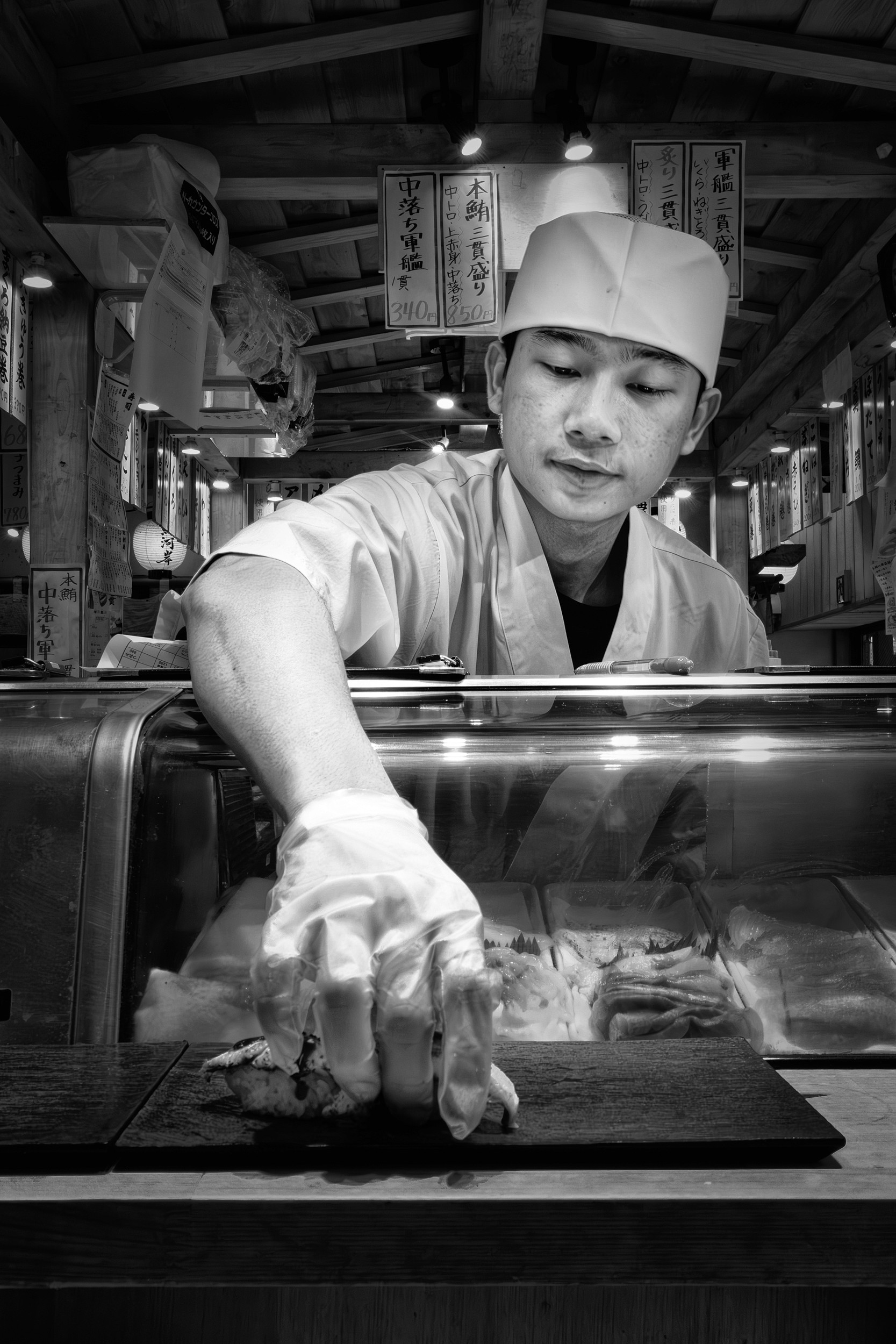 Black and white close-up image of a sushi chef reaching forward to place nigiri on a wooden board.