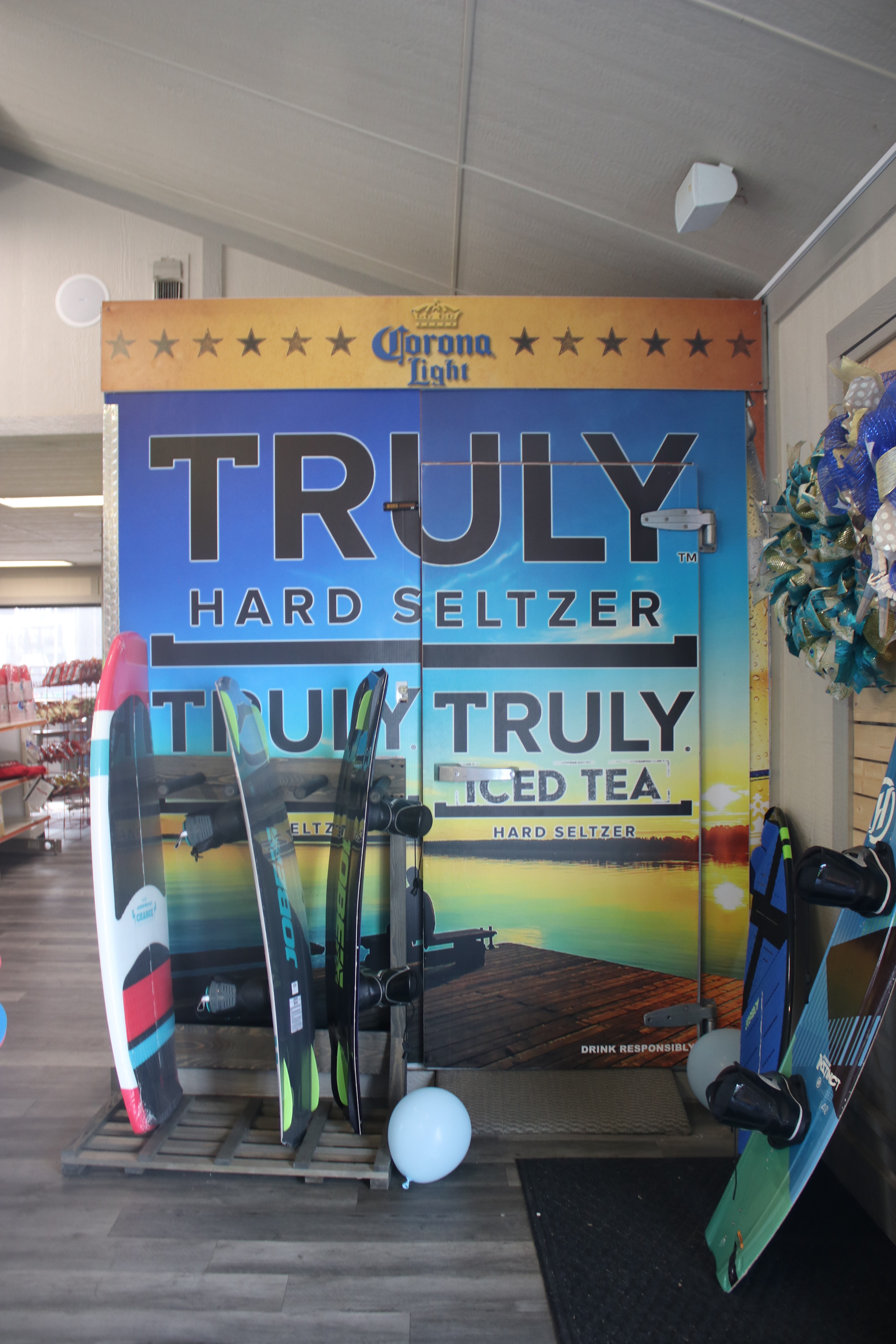 Surfboards are displayed on a wooden floor next to a vibrant advertisement for Truly Hard Seltzer and Iced Tea, with a picturesque sunset over a calm ocean in the background.