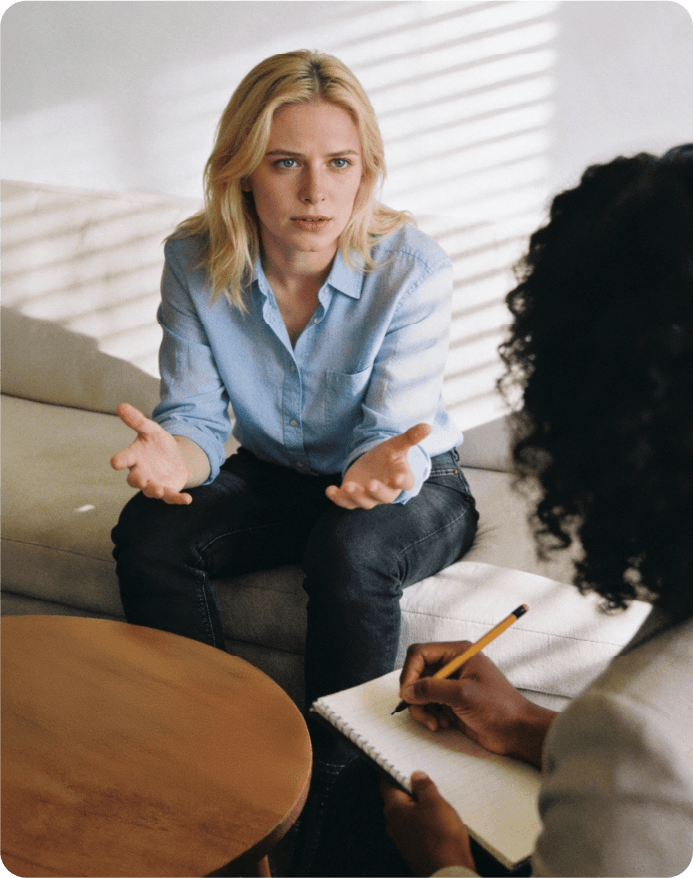 A blonde woman in a light blue button-down shirt and dark jeans sits on a light-coloured sofa, gesturing with her hands as she speaks while looking directly at the therapist. Her expression is serious and engaged. To her right, the back of another black female therapist's head, with dark curly hair, is visible, holding a notebook and pencil. In the foreground, a round wooden coffee table with nothing on it. The background features a white wall with blinds casting striped shadows. The lighting is soft and natural, creating a calm and intimate atmosphere.
