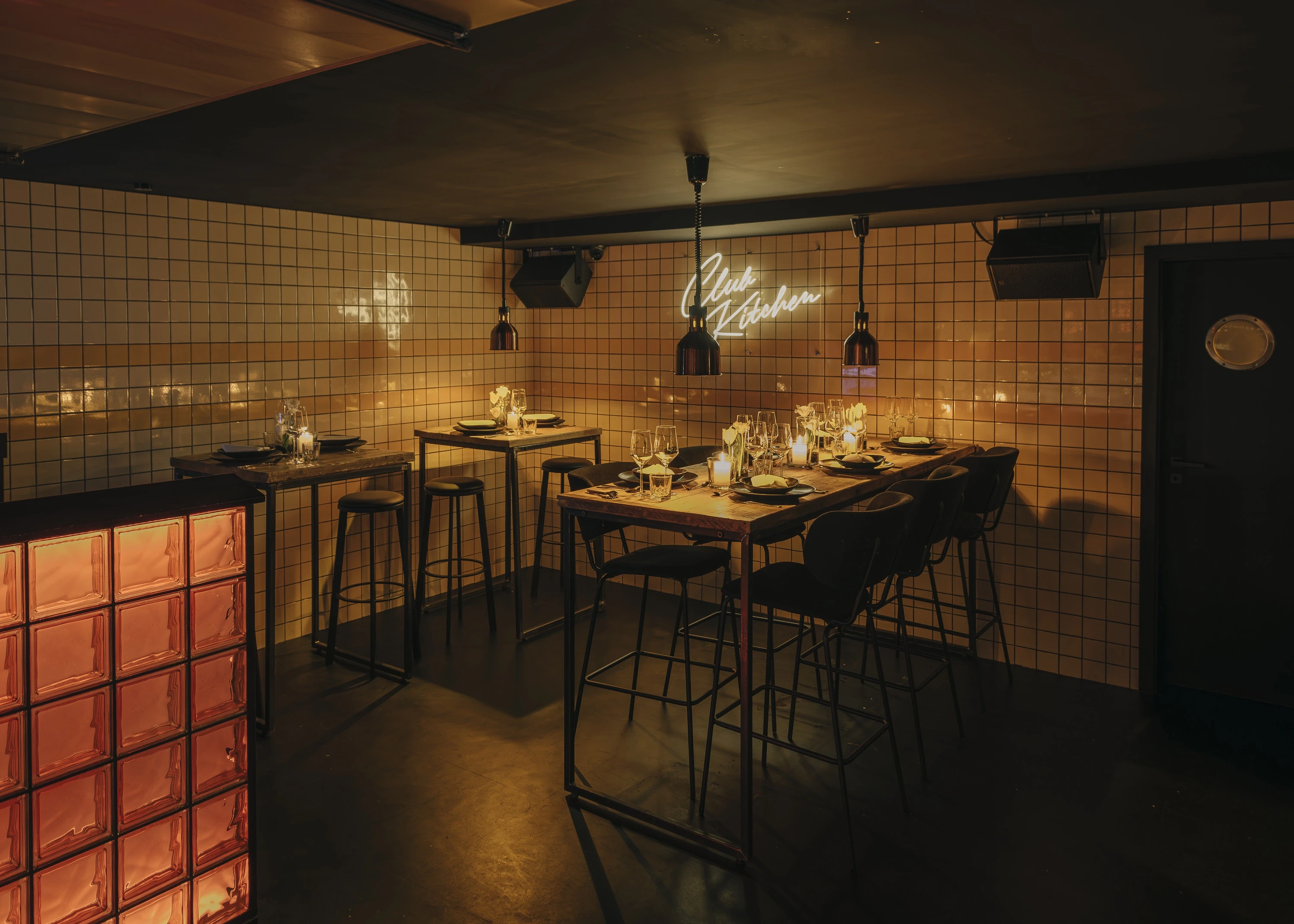 Dining corner in the Club Kitchen at NATE CLUB with beige tiled walls, black high tables set with candles and flowers, bar stools and warm pendant lights.