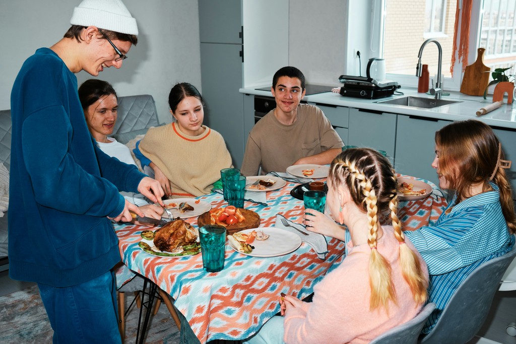 A group of friends enjoy a meal together in a cozy kitchen, with a young man carving a roast chicken at a table covered with a colorful patterned tablecloth, and everyone smiling and engaged in conversation.