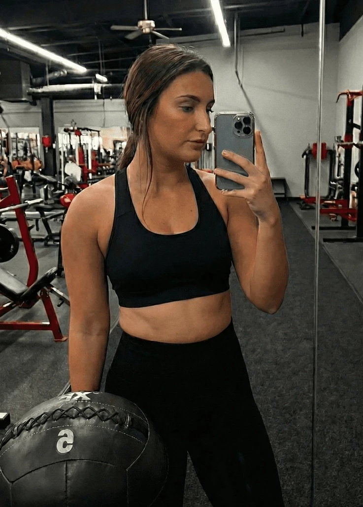 A person in athletic wear takes a mirror selfie in a gym, holding a medicine ball, surrounded by fitness equipment like weights and machines.