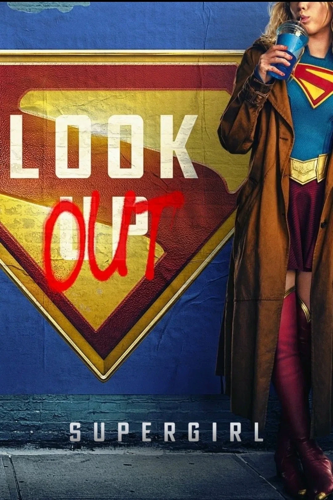 A woman dressed as a superhero stands beside a graffiti-covered wall featuring a large shield emblem, sipping from a blue cup; the background prominently displays the words "Look Out" and "Supergirl."