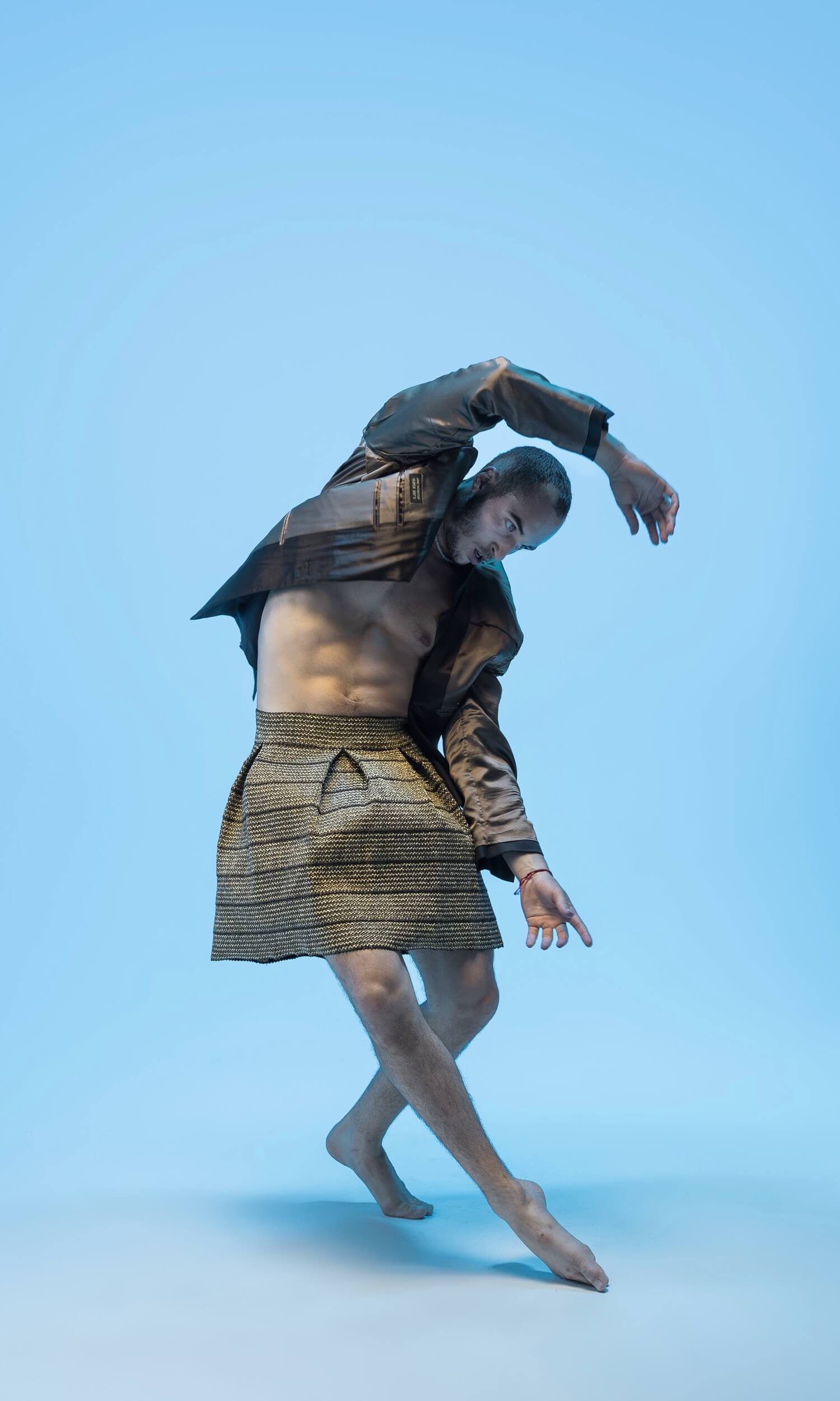 A circular studio portrait of dancer Alex Arce captured mid-leap in an expressive, dynamic contemporary dance pose against a light blue backdrop.