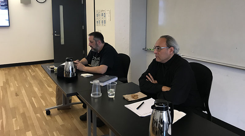 A photo of two men sitting at a desk and taking notes.