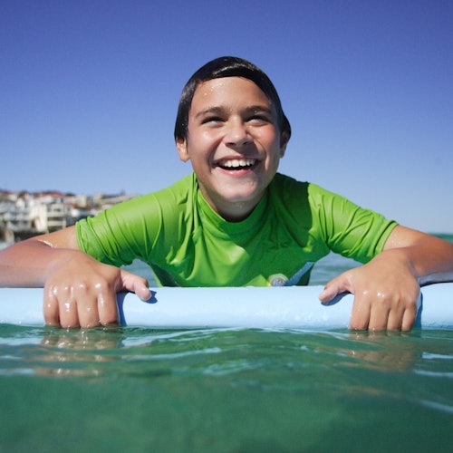 Ein lächelnder Junge in einem grünen Hemd hält eine blaue Poolnudel im Wasser, während im Hintergrund Gebäude sichtbar sind.