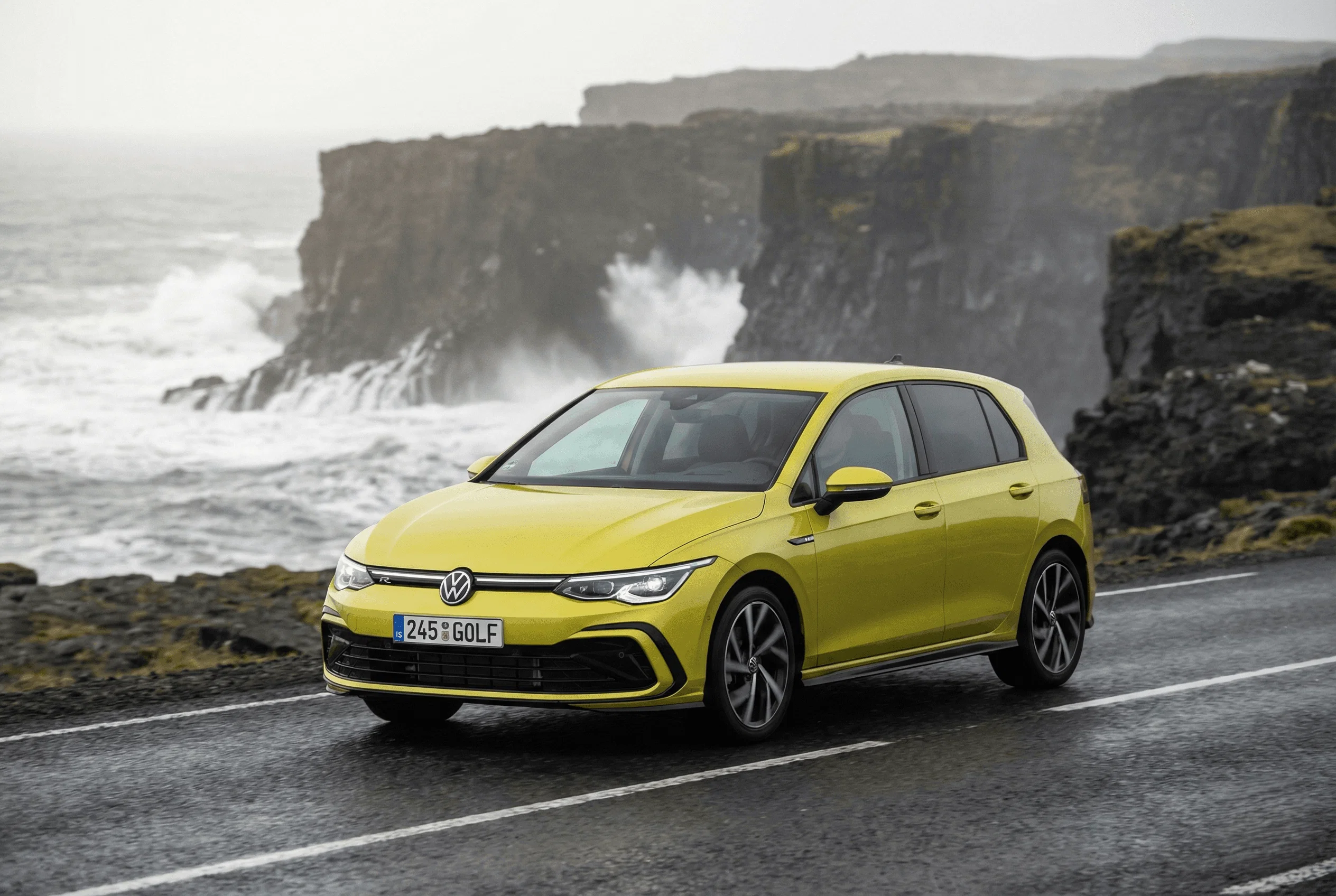 A yellow VW Golf parked near the edge of a cliff with ocean waves crashing against rocks.