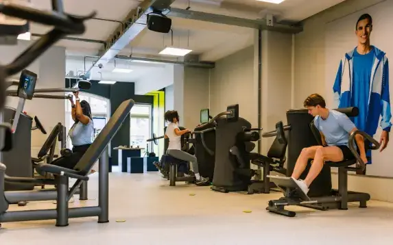 A person with curly hair sits on machine, watching a screen in a gym.