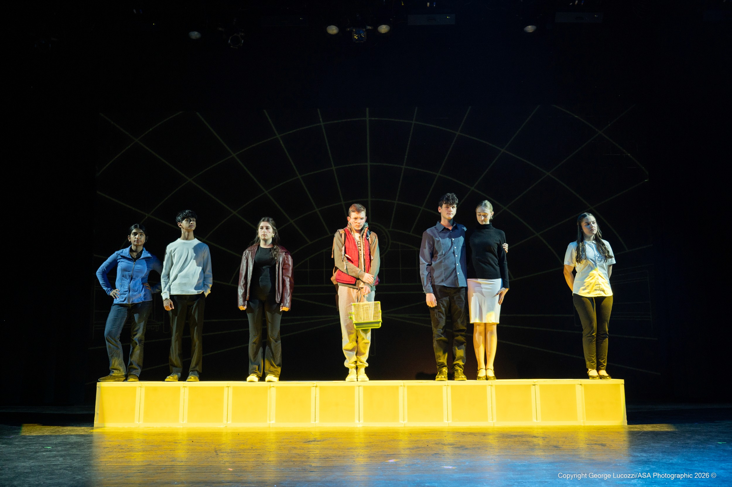 Christopher stands centered among six “Voices” atop a line of bright yellow-lit blocks, silhouetted against a projected outline of a train station, creating bold graphic contrast against the dark upstage wall.