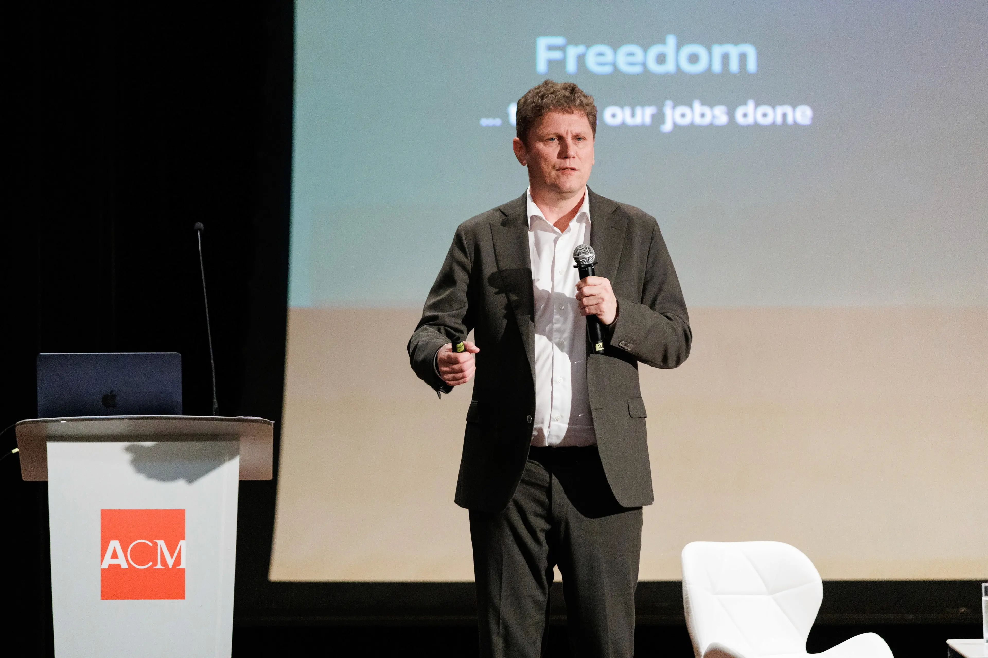 A speaker stands on stage, gesturing while presenting at a conference, with a screen behind them displaying text.