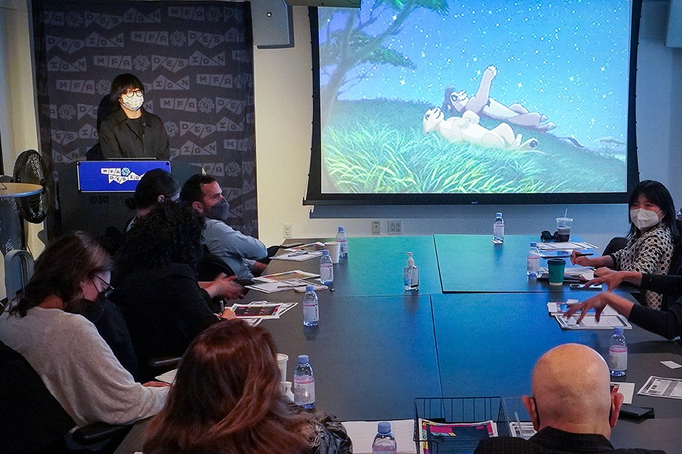A man dressed in black is giving a speech in front of an audience, and on the right side is an image projected of a cartoon still with two lions sitting on their back in the grass, looking at the starry sky