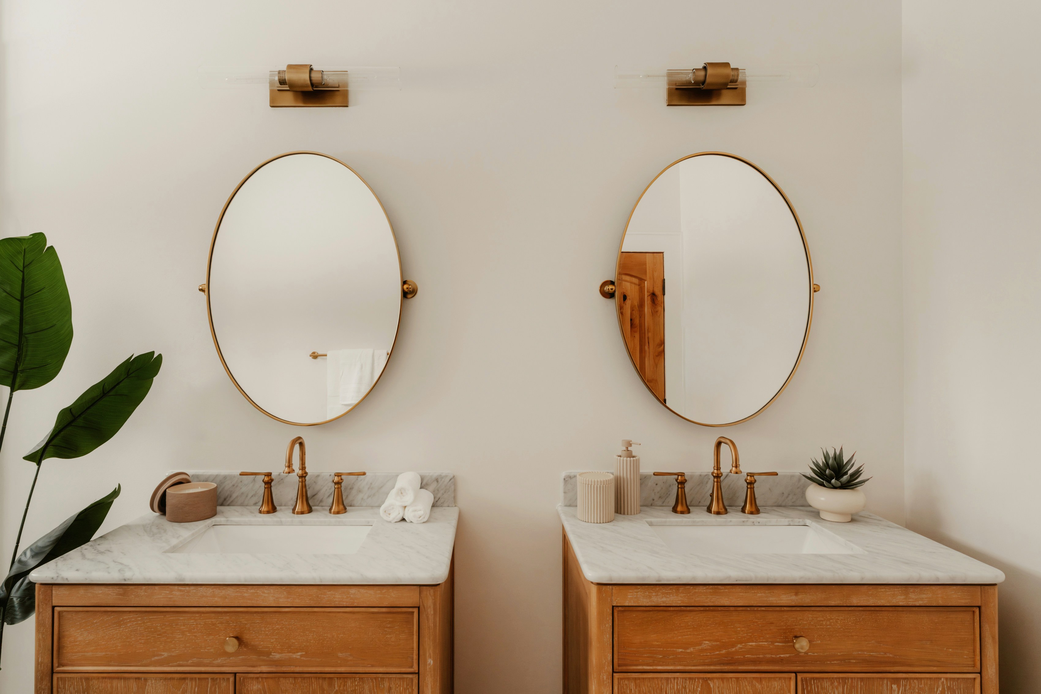 Two wooden vanities with oval mirrors and gold fixtures.