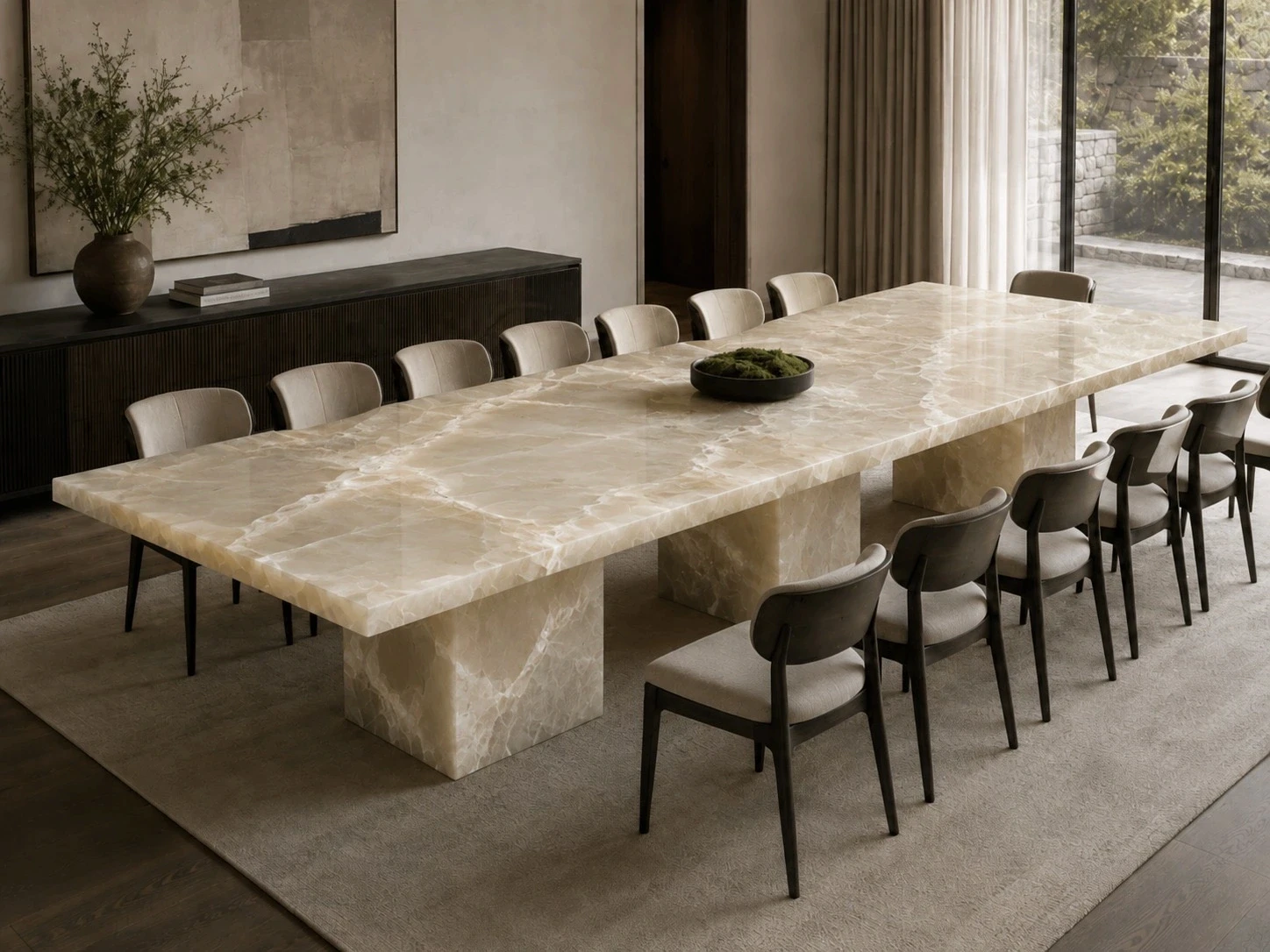 Long cream onyx dining table with a thick slab top and matching stone pedestal bases, surrounded by twelve taupe upholstered chairs with dark wood frames, on a textured neutral rug in a daylit dining room with a dark fluted sideboard, potted branches, abstract wall art, sheer curtains, and floor-to-ceiling windows overlooking a stone wall and greenery.