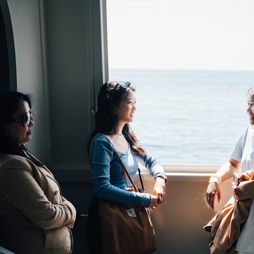 Tři ženy hovoří u okna s výhledem na moře za nimi, za přirozeného světla.