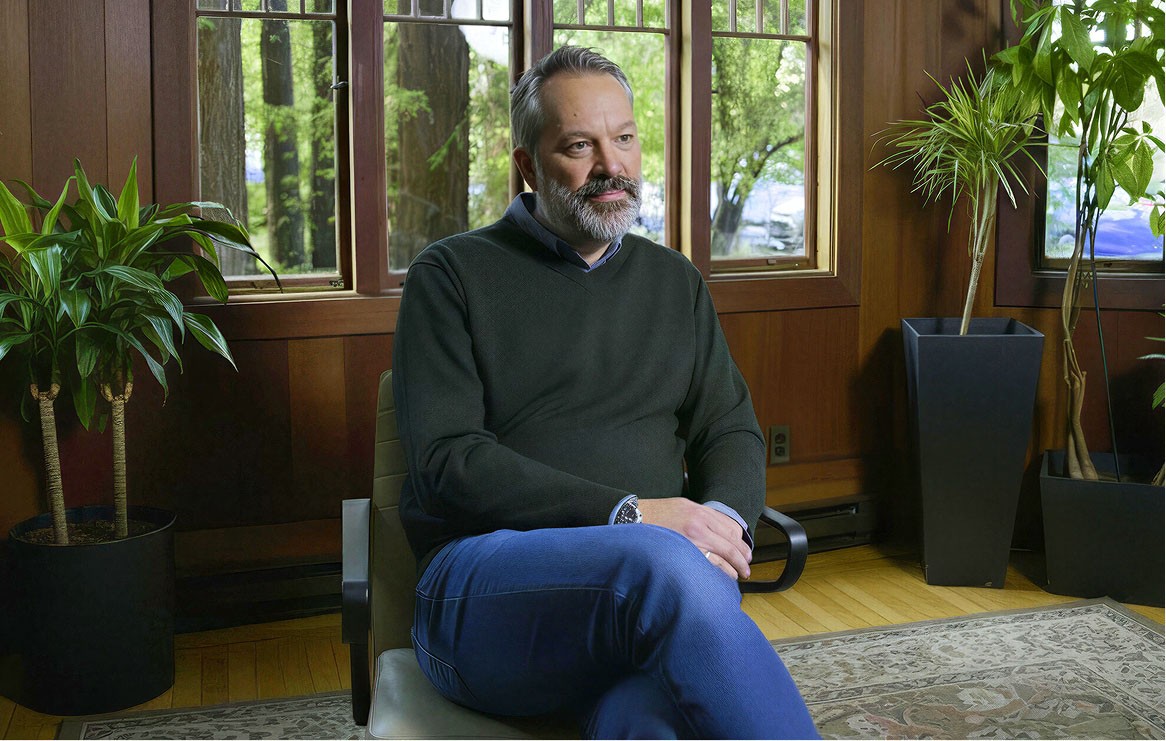Man sitting in a chair with a window and trees in the background looking relaxed.