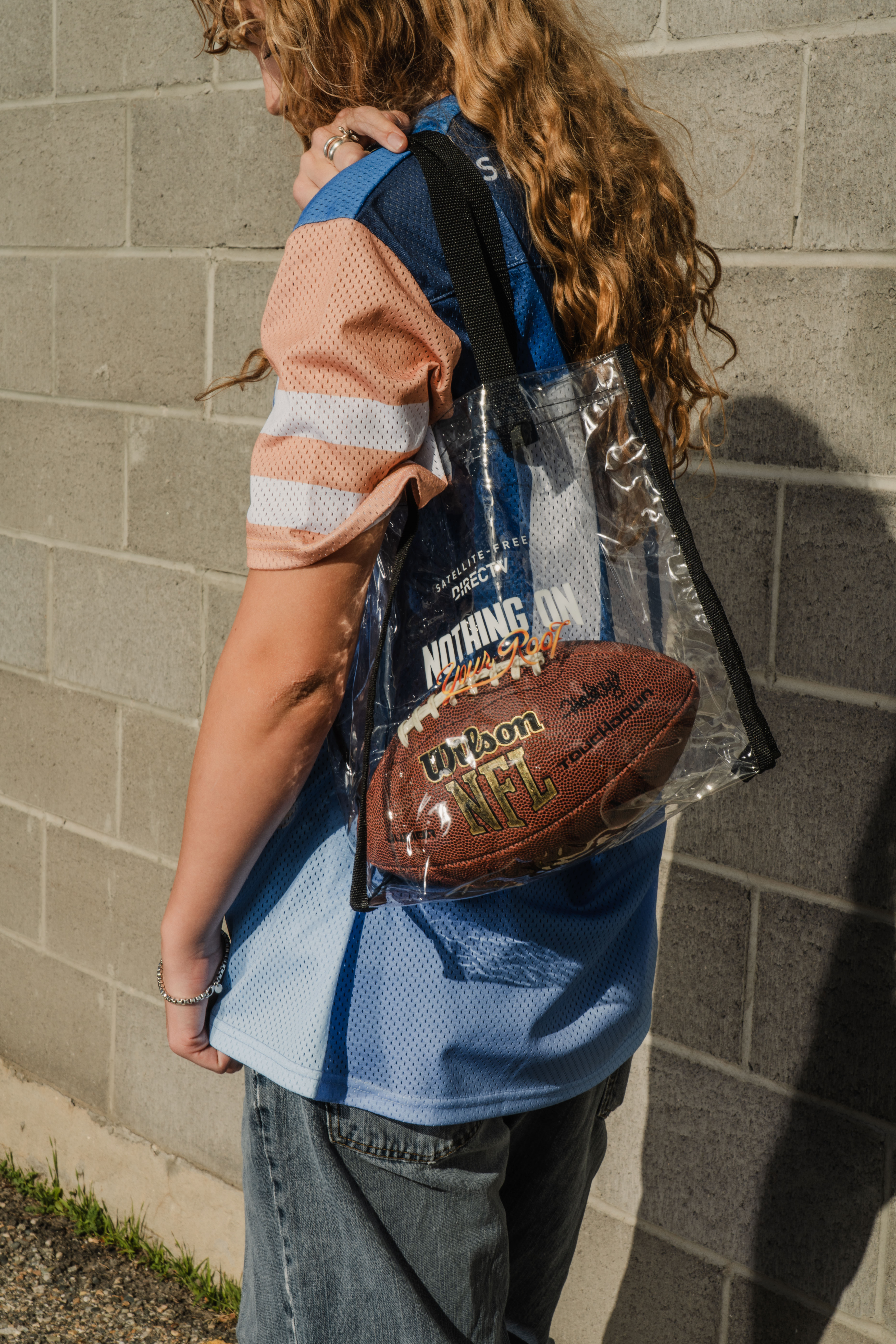 A girl next to a cinderblock wall looking to the left with long curly blond hair. She is wearing blue jeans, a custom football jersey, and a clear transparent stadium tote that has a football inside.
