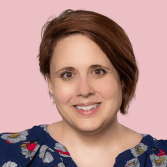 A smiling woman with short brown hair styled in a side-swept look wearing a navy blue top with colorful floral print, photographed against a soft pink background.