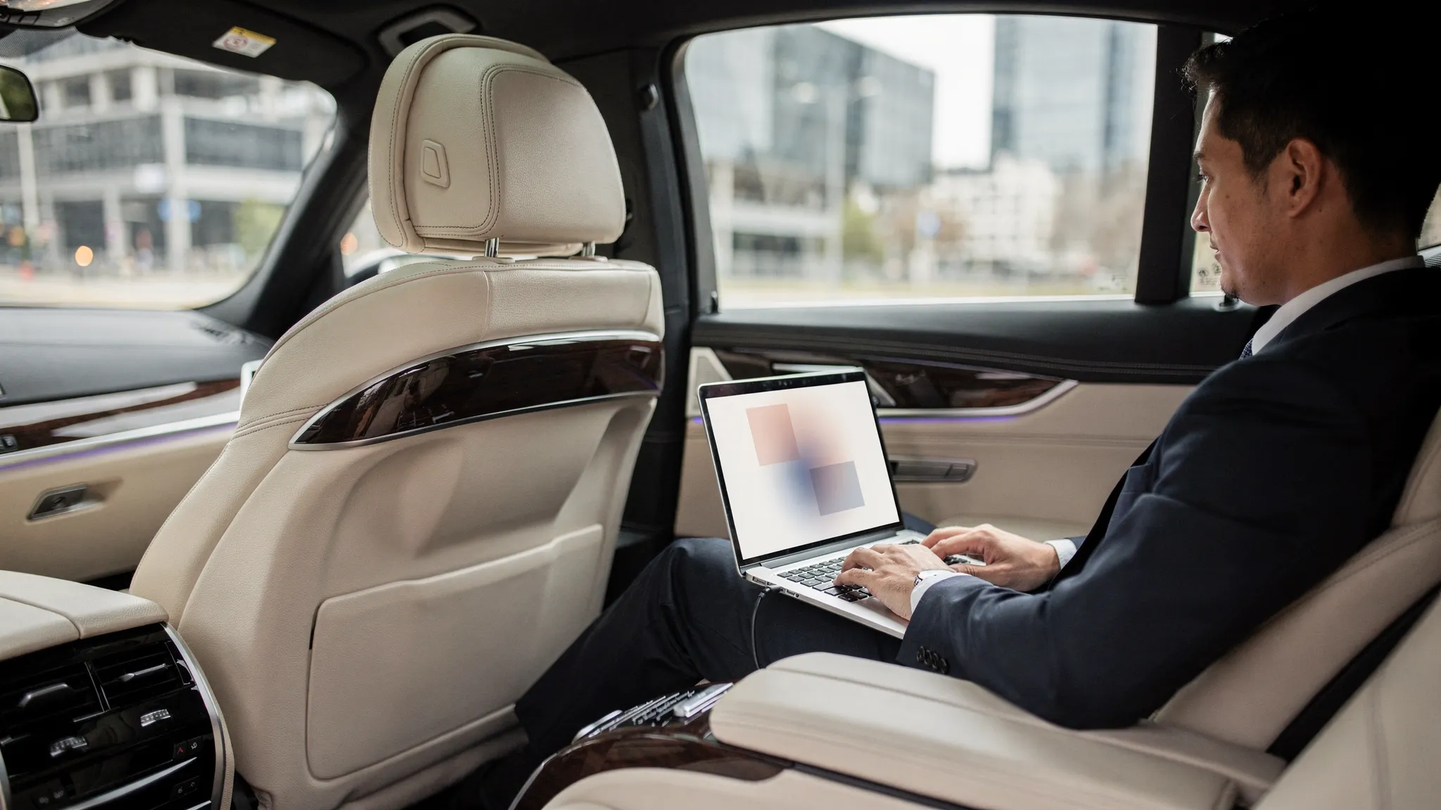 Interieur van een luxe auto met een passagier op de achterbank die rustig werkt met een laptop; het laptopscherm staat in de juiste richting en toont geen herkenbare data. Op de achtergrond is een zachte, onscherpe stadsroute zichtbaar door het raam.
