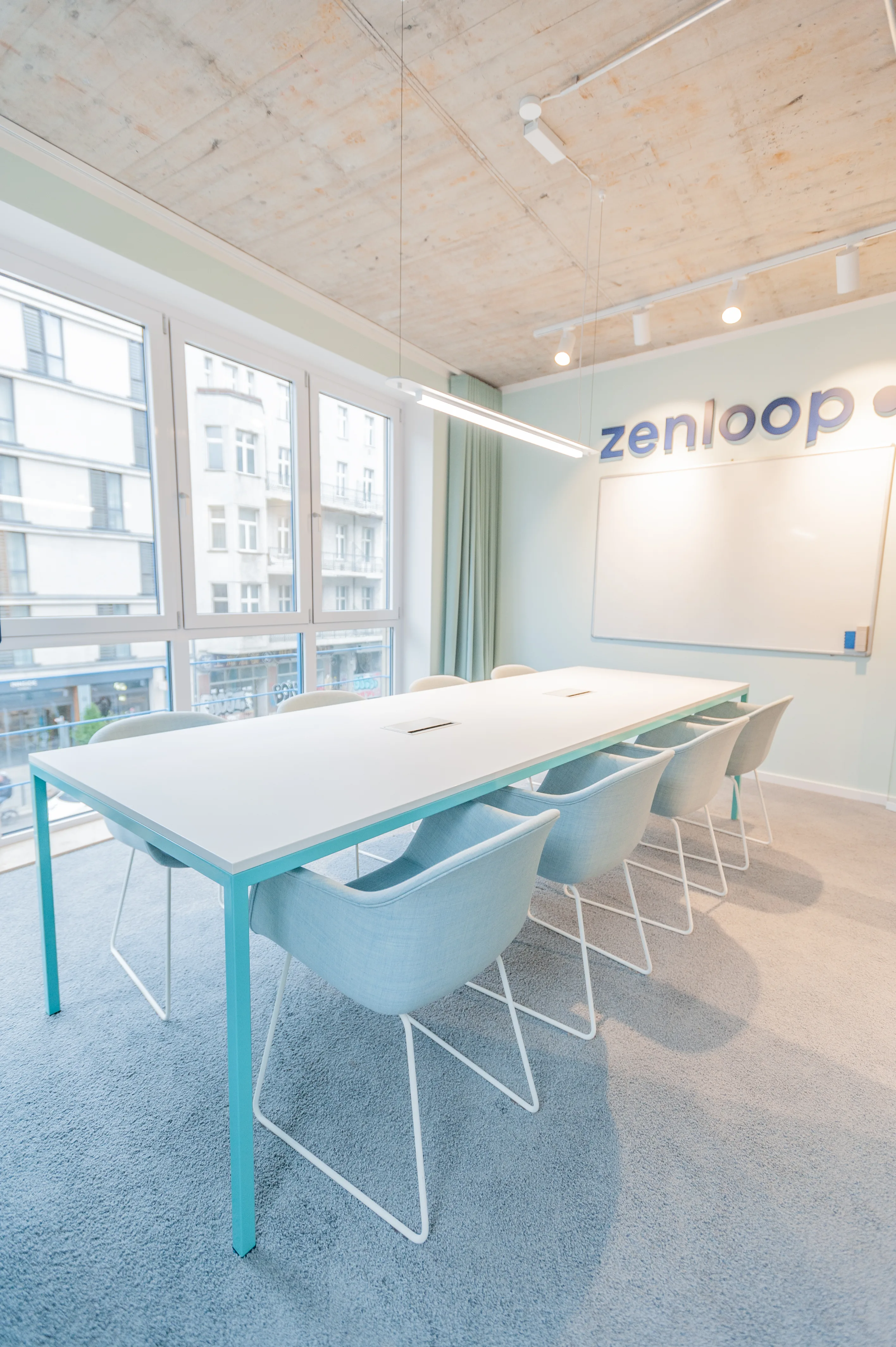 Mint-green conference room at ZENLOOP with a turquoise-framed white table, pale blue chairs, a whiteboard under the zenloop logo, green curtains and blue-grey carpet.