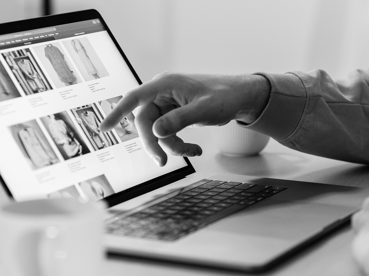 Black-and-white close-up of a person browsing product images on a tablet beside a laptop, with a hand hovering over the screen.