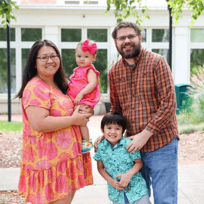 Alt tag: Portrait of Tricia Murphy with her family standing outdoors, smiling together in front of a building, with two young children held close by their parents