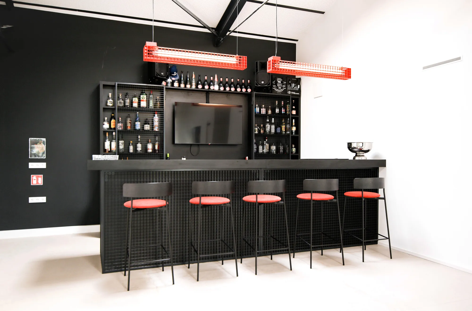 Black bar with red leather stools, TV and shelves of bottles against a dark wall.