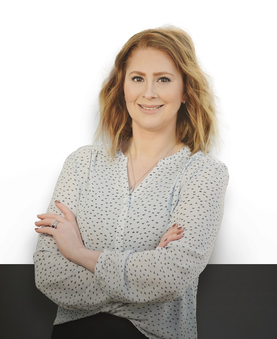 corporate headshot female with light skin and reddish blonde hair wearing a cream blouse with black spots, her arms crossed casually with a slight smile. 