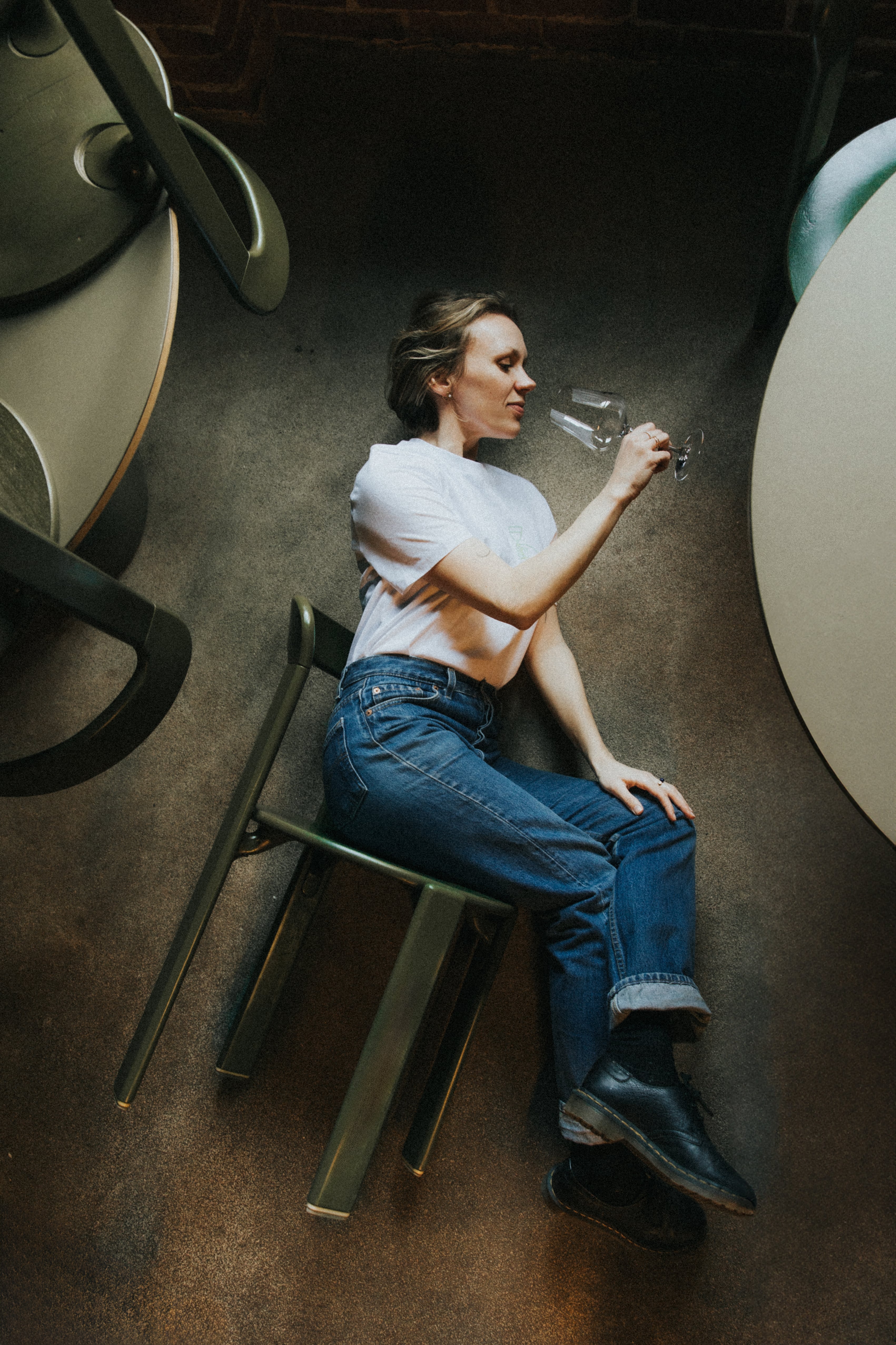 Kobieta w białej koszulce i dżinsach leży na szarej podłodze, trzymając kieliszek z winem. Opiera się o zielone krzesło ze spokojnym wyrazem twarzy.