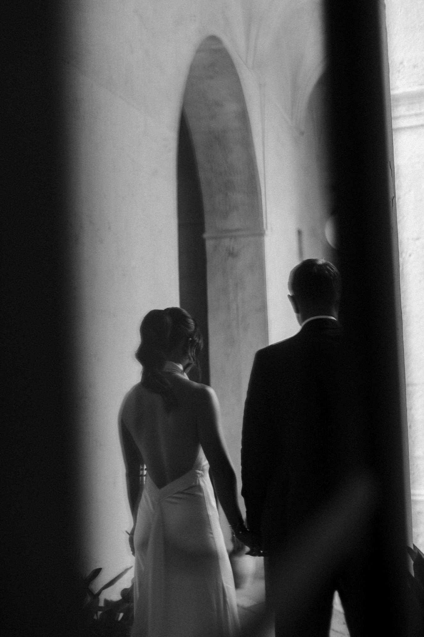 Black-and-white portrait of Toey and Otto from behind, holding hands beside an arched doorway